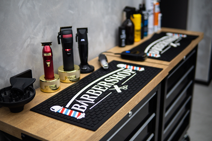 Elegancka mata z logo barbershopu, obok której starannie ułożono trzy maszynki do strzyżenia, suszarki oraz profesjonalne akcesoria fryzjerskie.