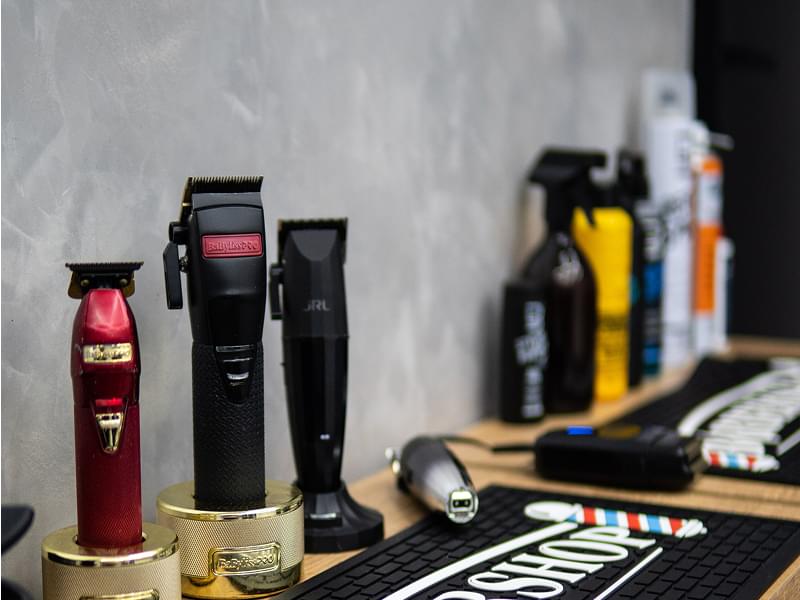 Blat na kółkach w salonie barberskim. Na czarnej silikonowej macie z napisem BARBERSHOP leżą: trzy maszynki (czerwona, czarna, czarna), złoty i czarny stojak na ostrza, nożyczki, trymer bezprzewodowy, mały czarny odkurzacz do włosów. Wszystko bardzo schludnie ułożone.