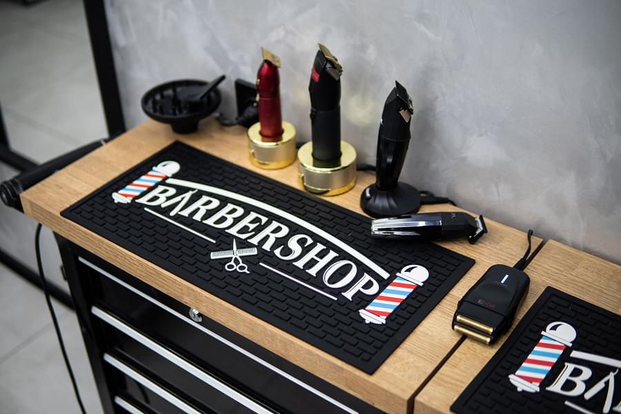 Bardzo zbliżone ujęcie blatu z maszynami do strzyżenia. Trzy główne maszynki: czerwona BaByliss gold z złotymi elementami, czarna BaByliss, czarna JRL. Przed nimi stoi srebrny trymer, w tle żółte, niebieskie i pomarańczowe butelki kosmetyków, czarna mata barberska z białym napisem.