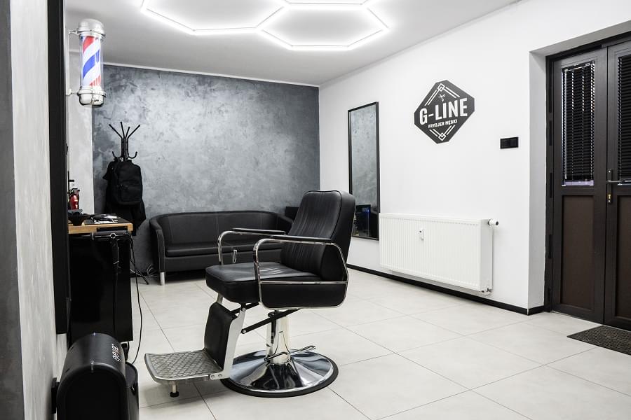 Szeroki kadr wnętrza salonu barberskiego. Centralnie czarny fotel fryzjerski z podnóżkiem, duże lustro z logo G-LINE, szara teksturowana ściana, umywalka z czarnym fotelem do mycia włosów po lewej, regały z kosmetykami, klasyczny barberski słupek, ciemne drzwi wejściowe, minimalistyczny i czysty wystrój.