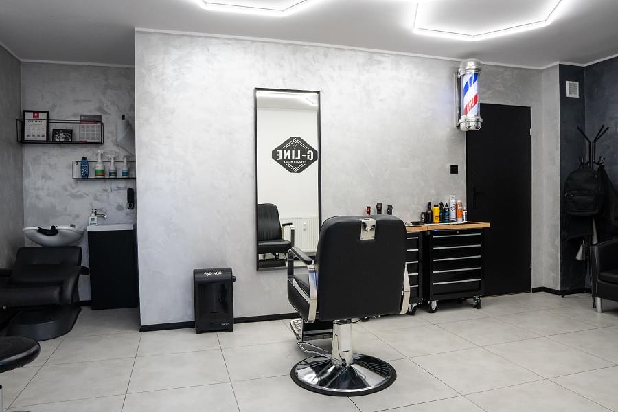 Nowoczesny, jasny salon barberski z szarą teksturowaną ścianą. Hexagonalne lampy LED na suficie, czarne skórzane fotel barberski z chromowanymi elementami na środku, duże prostokątne lustro, obok dwa czarne regały narzędziowe na kółkach, klasyczny biało-czerwono-niebieski słupek fryzjerski na ścianie, telewizor z logo G-LINE nad konsolą z laptopem.