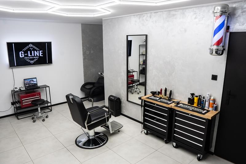Podobny kadr jak zdjęcie 3, nieco inny kąt. Czarny fotel barberski na lśniącej podłodze z kołami, duże lustro z logo G-LINE, szara ściana strukturalna, biało-czerwono-niebieski słupek fryzjerski, czarna sofa dwuosobowa w rogu, wieszak na ubrania, kaloryfer, ciemne drzwi, sufit z geometrycznymi LED-ami.