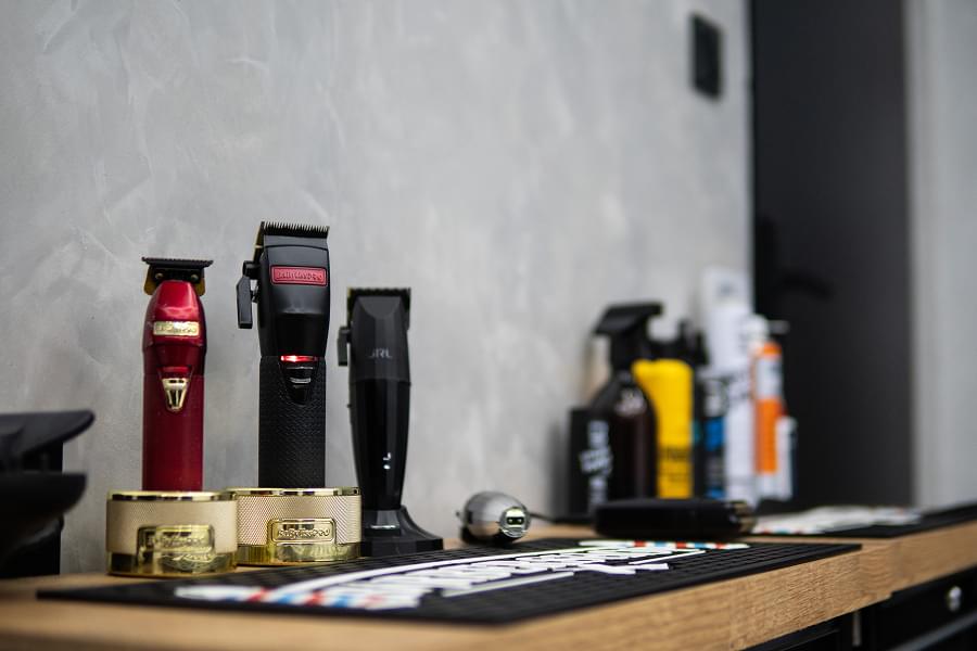 Bliski plan blatu roboczego w salonie barberskim. Na czarnym podkładzie z napisem BARBERSHOP stoją trzy maszynki do strzyżenia: czerwona BaByliss Gold, czarna BaByliss, czarna JRL. Obok stojaki na nożyce i trymer, butelki kosmetyków w tle, złote i czarne podstawki pod maszynki, kabelki zwisające.