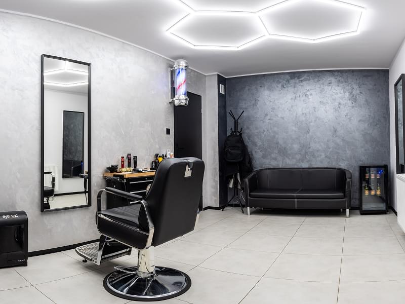 Salon od środka, na pierwszym planie czarny fotel do strzyzenia, który stoi przed lustrem a obok niego kosz na włosy i szafka z maszynkami oraz innymi akcesoriami do golenia. na Dalszym planie jest wieszak na kurtki, obok czarna kanapa oraz lodówka z napojami w środku na tle.